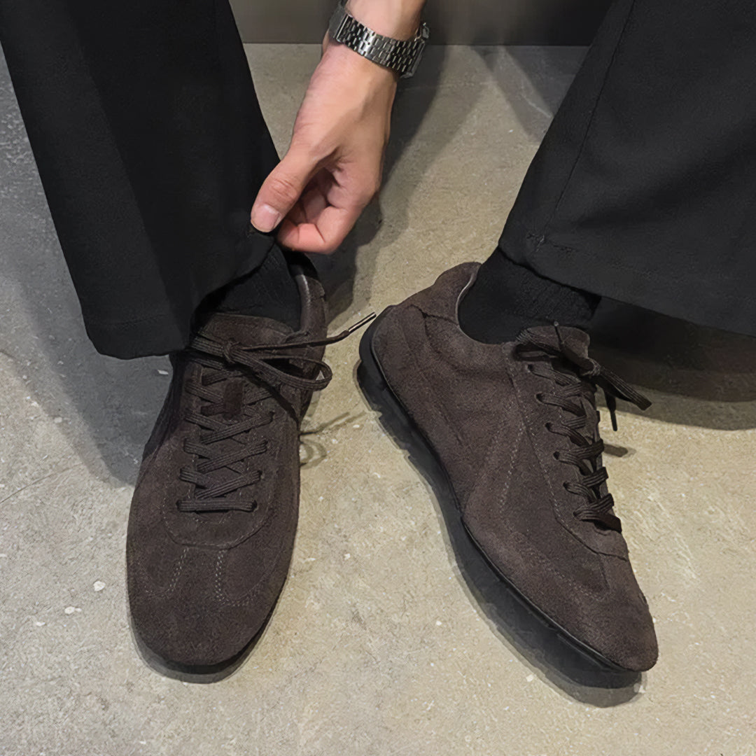 Brown suede sneakers worn with black pants on a concrete floor.