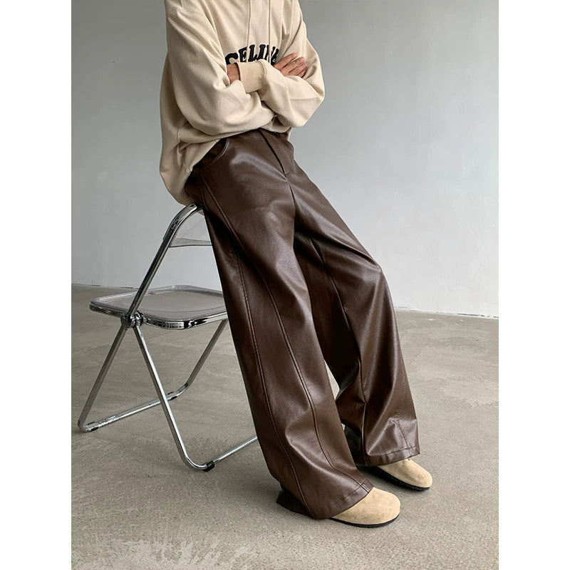Person wearing a beige sweatshirt and brown leather pants sitting on a metal chair against a plain wall.